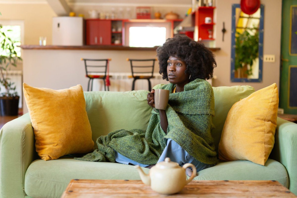 A woman wrapped in a green blanket sits on a couch, holding a mug and looking off to the side