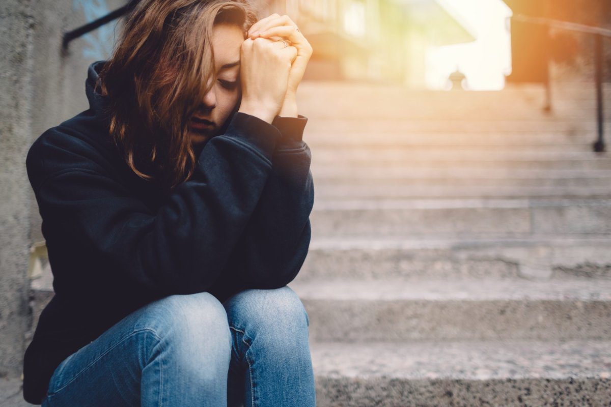 A sad-looking young person in a hoodie sits alone on steps with their head in their hands
